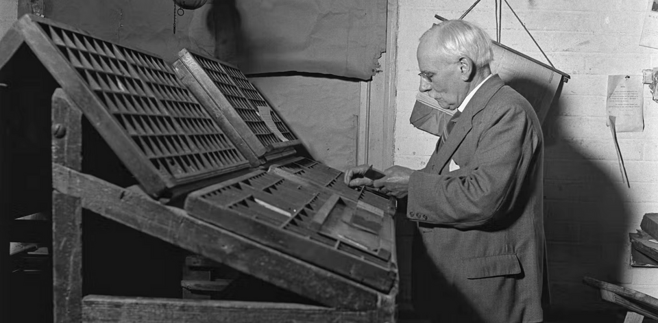 Black-and-white image of an older gentleman standing before some sort of contraption for organizing literal typefaces.